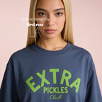 A woman wearing a navy blue crewneck sweatshirt with a bright green graphic that reads 'EXTRA PICKLES Club' on the front, set against a solid pale pink background. Original design by first row LA.
