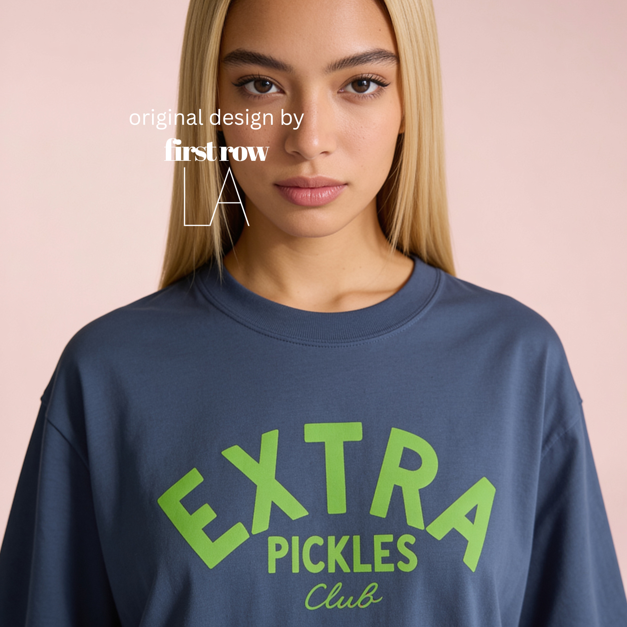 A woman wearing a navy blue crewneck sweatshirt with a bright green graphic that reads 'EXTRA PICKLES Club' on the front, set against a solid pale pink background. Original design by first row LA.
