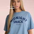 A blonde woman wearing a light blue oversized t-shirt with 'MIDNIGHT SNACK Society' printed in navy blue on the front. Original design by first row LA.