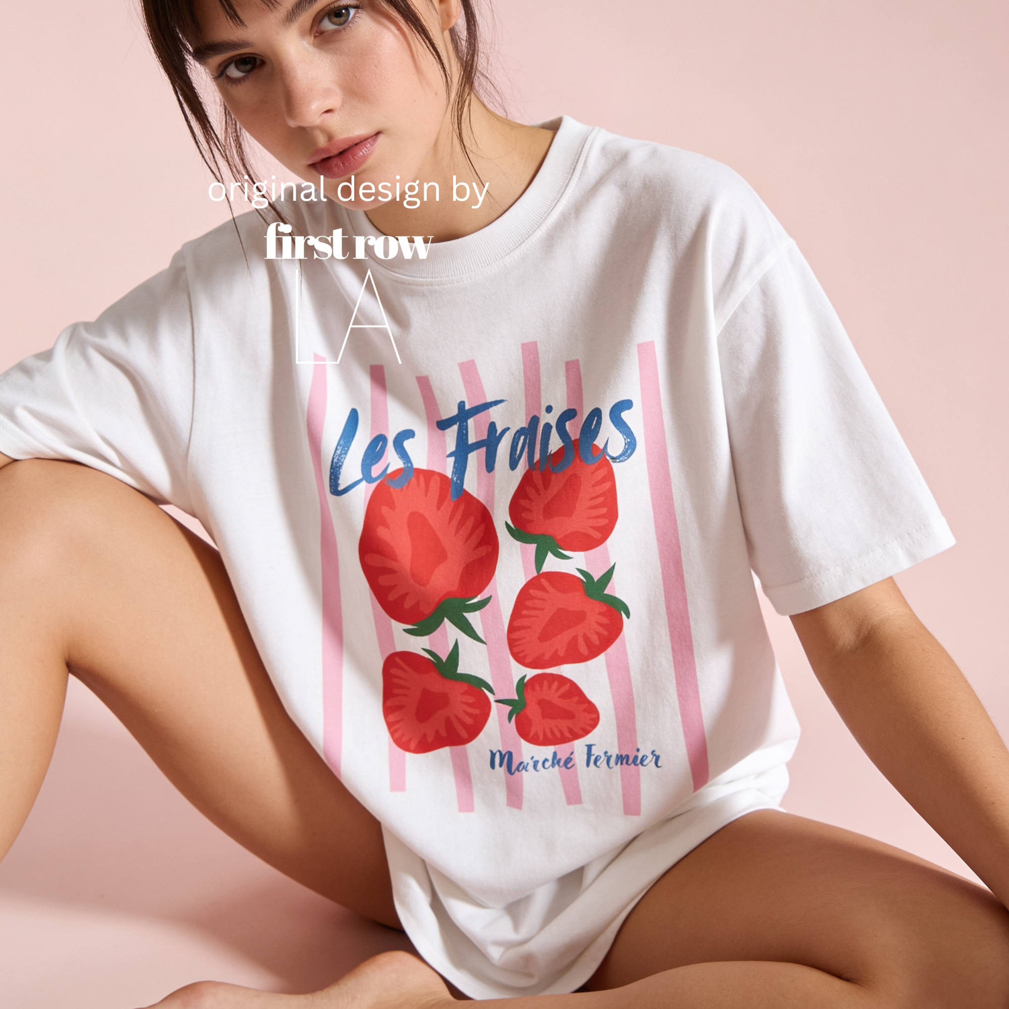 A woman wearing an oversized white graphic T-shirt with pink vertical stripes, red strawberries, and blue script text reading 'Les Fraises' and 'Marche Fermier', posed against a soft pink studio background. Original design by first row LA.