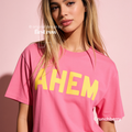 A woman wearing a bright pink T-shirt with the word 'AHEM' printed across the chest in large yellow letters.

Original design and copyright by first row LA, August 4th 2026.