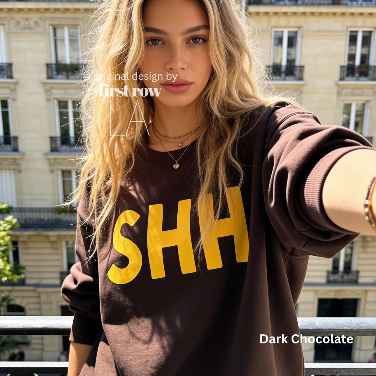 A woman wearing a dark chocolate brown sweatshirt with the letters 'SHH' printed in bold yellow across the chest. 

Original design and copyright by first row LA, August 4th 2026.