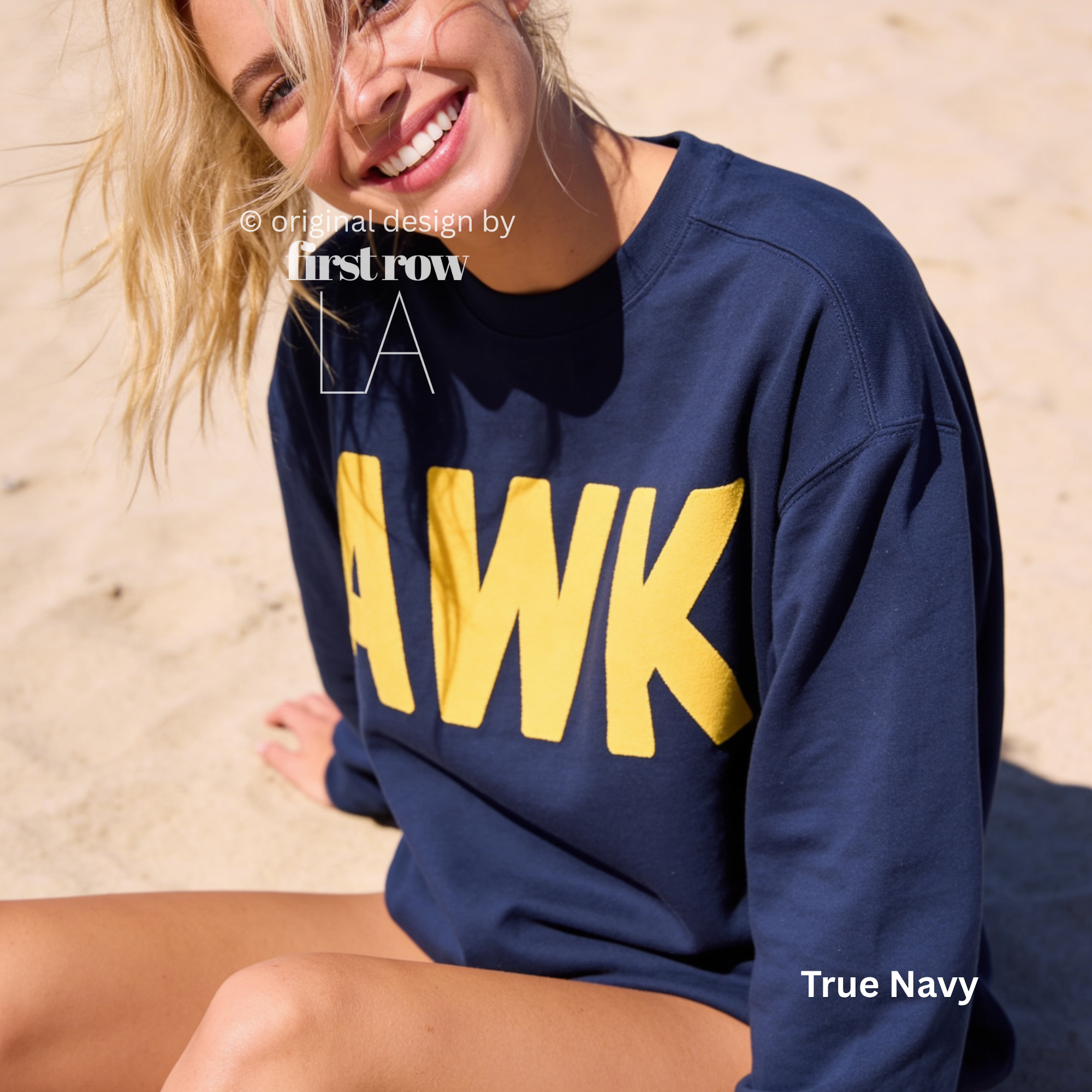 A woman sitting on a sandy beach wearing a navy blue sweatshirt with large yellow 'AWK' lettering across the front. 

Original design and copyright by first row LA, August 4th 2026.