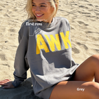 A woman sitting on a sandy beach wearing a grey long-sleeve sweatshirt with bold yellow 'AWK' lettering on the front. 

Original design and copyright by first row LA, August 4th 2026.