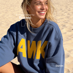A woman smiling on a sandy beach wearing a denim blue sweatshirt with large yellow 'AWK' lettering on the front. 

Original design and copyright by first row LA, August 4th 2026.