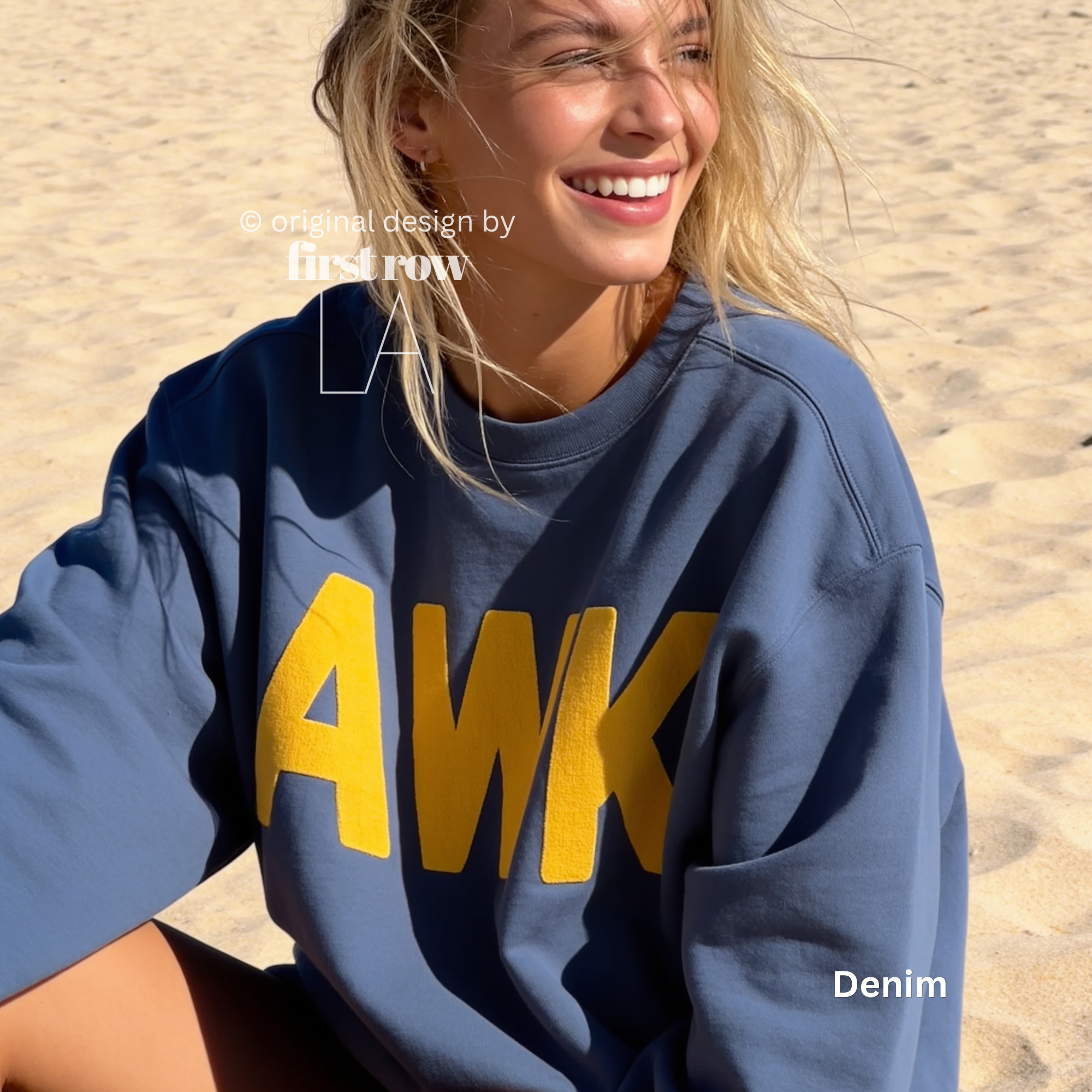 A woman smiling on a sandy beach wearing a denim blue sweatshirt with large yellow 'AWK' lettering on the front. 

Original design and copyright by first row LA, August 4th 2026.