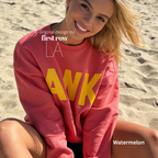 A woman sitting on a sandy beach wearing a coral pink sweatshirt with large yellow block letters spelling 'AWK' across the front. 

Original design and copyright by first row LA, August 4th 2026.