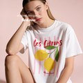 A woman wearing a white graphic T-shirt with pink vertical stripes, red 'Les Citrons, Marcher Fermier' lettering, and yellow lemon illustrations.

Original design and copyright by first row LA 2025.