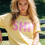 A woman wearing a pale yellow oversized T-shirt with large pink block letters spelling 'SHHH'. 

Original design and copyright by first row LA, August 4th 2026.