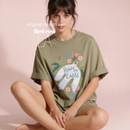 A woman sits wearing an oversized olive green T-shirt with a large front graphic of a white dove, flowers, and the words 'Peace on Earth.' 

Original design by first row LA.