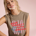 A woman wearing a sleeveless cream-colored t-shirt with a graphic print that reads 'POUR YOURSELF A CUP OF AMBITION' in red, decorated with pink vertical stripes, a blue moka pot, and a small coffee cup. 

Original design and copyright by first row LA.