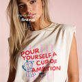 A woman wearing a sleeveless cream-colored t-shirt with a graphic print that reads 'POUR YOURSELF A CUP OF AMBITION' in red, decorated with pink vertical stripes, a blue moka pot, and a small coffee cup. 

Original design and copyright by first row LA.