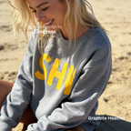 A woman wearing a heather gray sweatshirt with large yellow block letters spelling 'SHH'. 

Original design and copyright by first row LA, August 4th 2026.